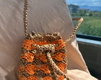 Könnte beinhalten: Handgefertigte Bucket Bag mit gewebtem Design. Die Tasche hat einen Kordelzugverschluss und einen langen Riemen. Der Körper der Tasche besteht aus einer Kombination aus beigem und orangefarbenem gewebtem Material. Die Tasche liegt auf einem hellrosa Stoff.