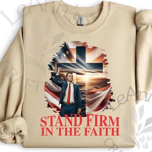 May include: A tan sweatshirt featuring a graphic of a man in a suit with an American flag and a cross. The text "STAND FIRM IN THE FAITH" is printed in red at the bottom of the design. The sweatshirt has long sleeves and a crew neck.