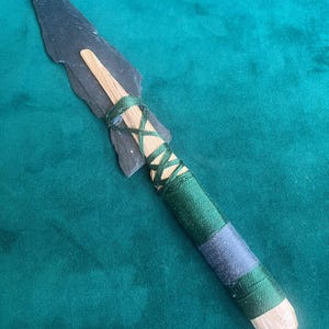 May include: A spear featuring a sharp, dark gray stone blade attached to a light wooden shaft. The connection point is secured with crisscrossed dark green thread, and the handle is wrapped in layers of dark green and gray thread.