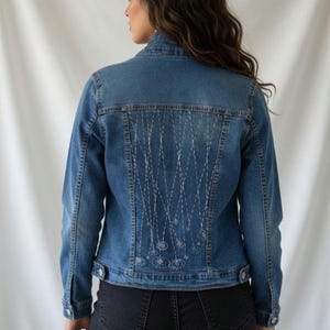 Könnte beinhalten: Blaue Jeansjacke mit weißer Stickerei auf dem Rücken. Die Jacke hat ein klassisches Knopfdesign mit langen Ärmeln und einem Kragen. Die Stickerei zeigt verbundene Kreise und Linien.