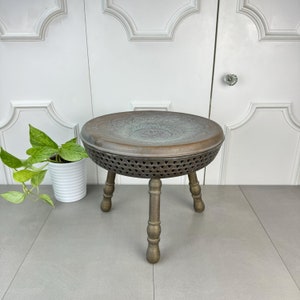 May include: A round, three-legged stool made of metal with a decorative, embossed top. The stool is brown and has a distressed finish.