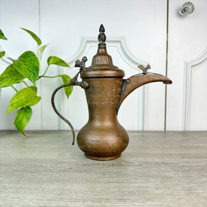 May include: An antique copper Arabic coffee pot with intricate engravings and a spout with a bird finial. The pot has a handle and a lid with a finial.