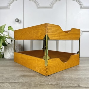 May include: A two-tier wooden document tray featuring a light brown oak finish with visible wood grain. The two trays are connected by vertical brass-colored metal corner brackets, and each tray has a curved cutout on the front edge.