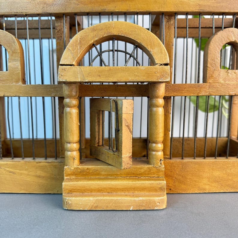 May include: A wooden birdcage with a small door that is open. The cage has a decorative arched top and a small porch with columns.