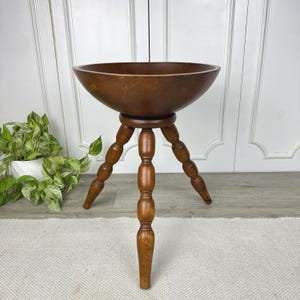 May include: A vintage wooden bowl on a tripod stand. The bowl is a deep brown color and has a wide, shallow shape. The stand has three legs and a central support, all made of the same wood. The piece is likely used for decorative purposes.