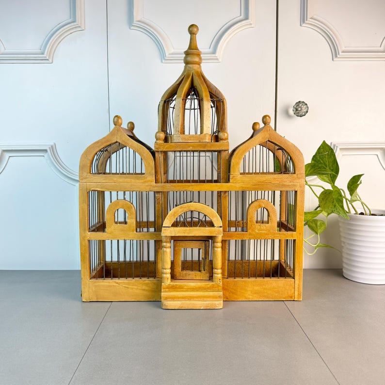 May include: A wooden birdcage with a domed top and intricate details. The cage has multiple compartments and is made of natural wood with a light brown finish.