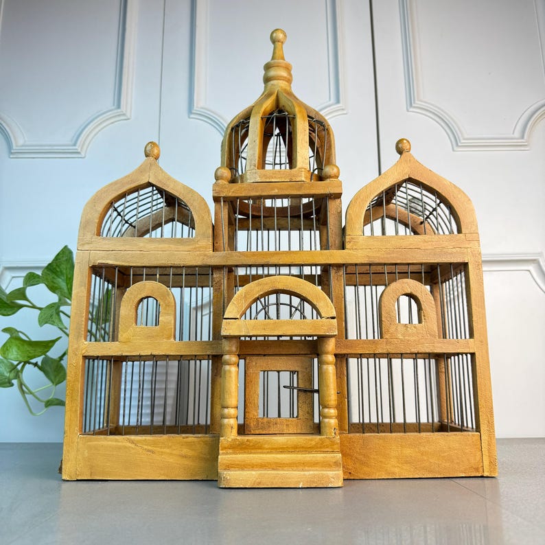 May include: A large, wooden birdcage with a domed top and intricate details. The cage is made of natural wood and has a wire mesh interior. The cage has a small door on the front and is decorated with decorative accents.