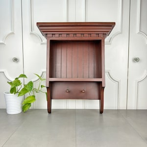 May include: A dark brown wooden wall shelf with a small cabinet and two knobs. The shelf has a rectangular top with decorative molding and a vertical slatted back. The shelf is approximately 24 inches tall.