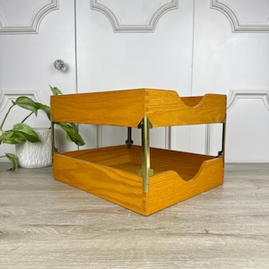 May include: A two-tier wooden document tray featuring a warm honey-toned finish with visible wood grain. The structure is supported by four vertical brass-colored metal corner brackets that connect the top and bottom trays, each with a cutout.