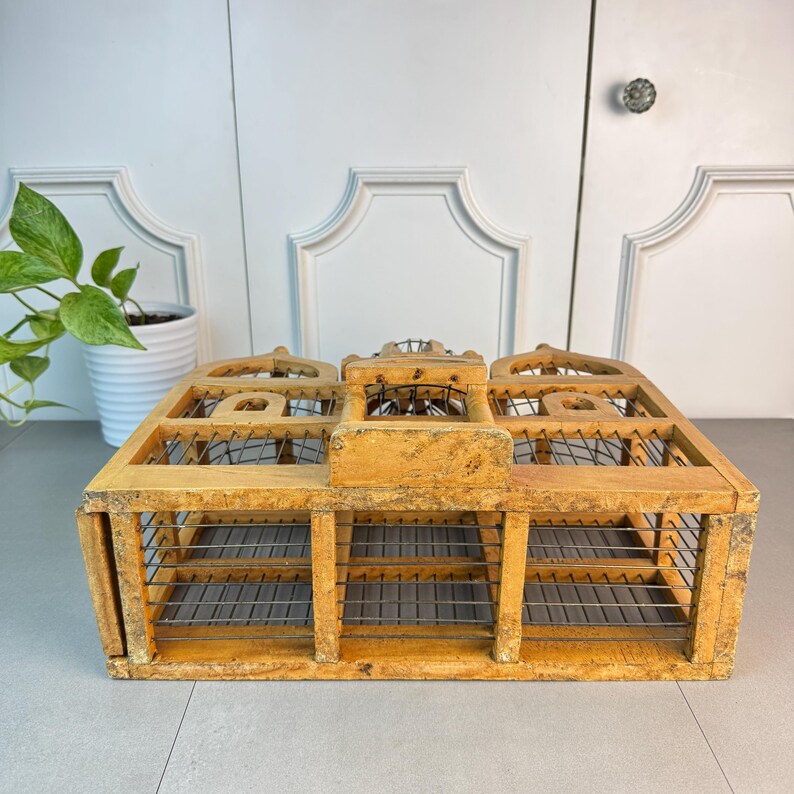 May include: A wooden birdcage with a decorative, ornate design. The cage is made of light brown wood and has a wire mesh bottom. The cage is empty.