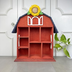May include: Red wooden barn-shaped shelf with a sunflower and "Moo Moos" sign on top. The shelf has six compartments.