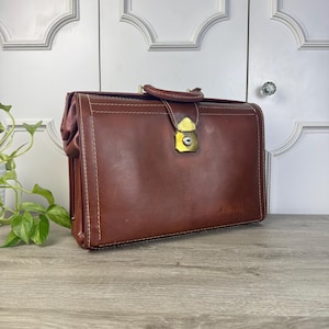 May include: A brown leather briefcase with a brass clasp and handle. The briefcase has white stitching and a rectangular shape. The bag is sitting on a wooden surface.