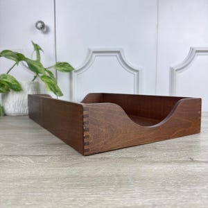 May include: A rectangular wooden desk tray with a medium brown finish, featuring visible finger joint construction at the corners and a curved cutout on the front panel for easy access to documents or items stored within the open-top container.