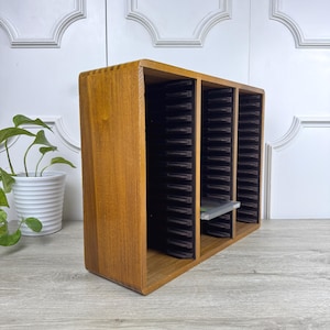 May include: A wooden CD storage rack with three vertical compartments. The rack is made of light brown wood and has a rectangular shape. Each compartment holds multiple CD cases. The rack is designed to hold CDs and keep them organized.