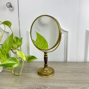 May include: A vintage-style, round vanity mirror with a gold-toned frame and base. The mirror has a dark brown wooden stem and a decorative, ornate base. The mirror is designed to sit on a table or vanity. The mirror is approximately 10 inches tall.