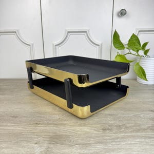 May include: A two-tiered gold metal desk organizer with black trays. The organizer is made of metal with a gold finish and has black plastic handles.