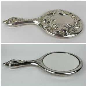 May include: A silver-toned hand mirror with an oval mirror surface and a decorative handle. The handle features floral and scrollwork designs. The back of the mirror has a similar decorative pattern. The mirror is likely vintage or antique.