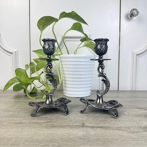 May include: A pair of ornate, silver-toned candle holders. Each holder features a detailed floral design on the cup and base, with a curved stem. The holders are displayed on a wooden surface, with a white pot and green plant in the background.