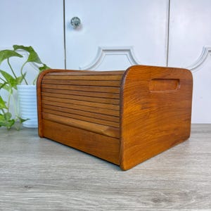 May include: A wooden bread box with a roll-top lid and a handle on the side. The box is made of light brown wood and has a curved top. The bread box is designed to keep bread fresh and is a kitchen storage item.