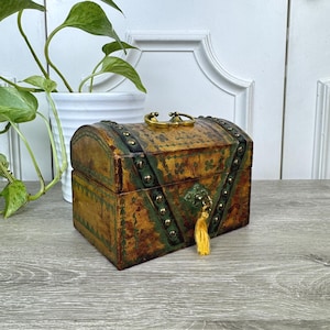 May include: A small, antique-style wooden chest with a curved top. The chest is painted in shades of brown and green, with decorative metal accents and a gold-colored handle. A yellow tassel hangs from the lock.