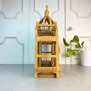 May include: A tall, wooden birdcage with a decorative, pointed roof. The cage is made of natural wood with a light brown finish and has a wire mesh interior. The cage is standing on a white surface.