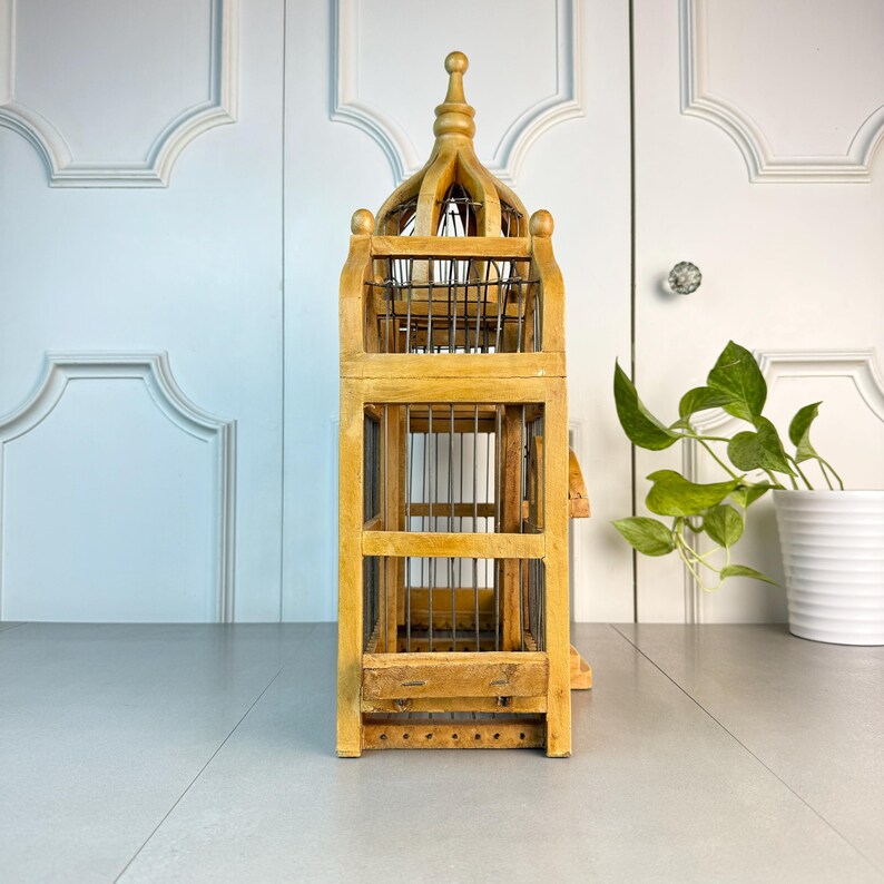 May include: A tall, wooden birdcage with a decorative, pointed roof. The cage is made of natural wood with a light brown finish and has a wire mesh interior. The cage is standing on a white surface.