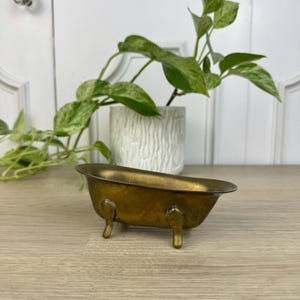 May include: A small, vintage-style brass bathtub dish with a smooth, metallic finish and four decorative claw-style feet. The basin has an elongated, oval shape with a wide, curved rim, showcasing a warm, aged golden-brown patina throughout.