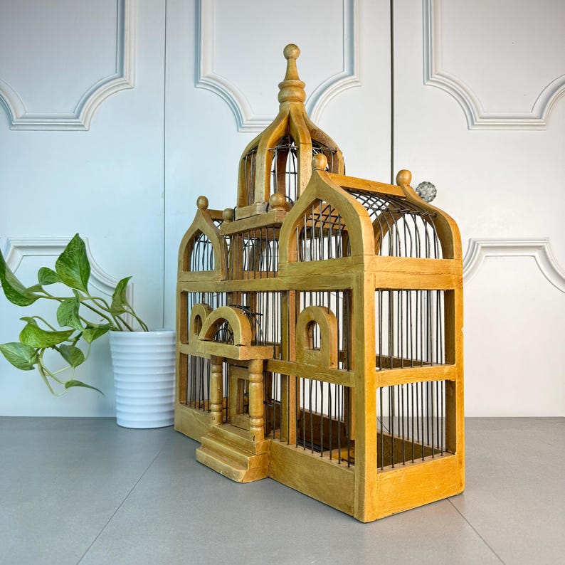 May include: A large, wooden birdcage with a decorative, ornate design. The cage is made of natural wood and has a light brown finish. The cage has a domed top and multiple levels. The cage is empty.
