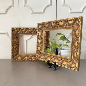 May include: Two ornate, gold-colored picture frames with detailed scrollwork. One frame is propped up, revealing a glimpse of a plant in a white pot. The frames are empty, ready for photos or artwork.