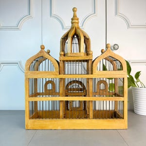 May include: A large, wooden birdcage with a domed top and intricate details. The cage is made of natural wood and has a wire mesh interior. The cage is empty and ready for a feathered friend.