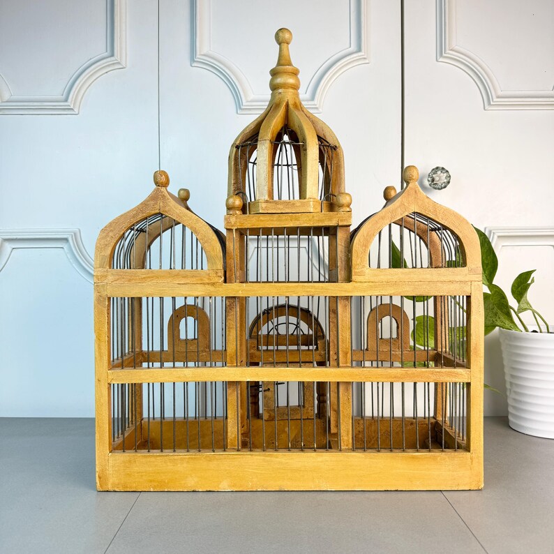 May include: A large, wooden birdcage with a domed top and intricate details. The cage is made of natural wood and has a wire mesh interior. The cage is empty and ready for a feathered friend.