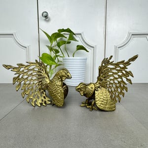 May include: Two antique brass bird sculptures with outstretched wings. The birds are detailed with feathers and stand on a gray surface. A white potted plant is in the background.