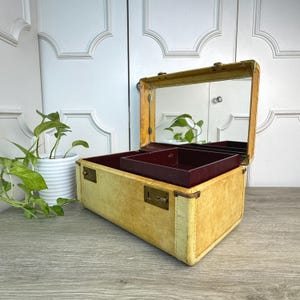 May include: A vintage yellow jewelry box with a rectangular shape, featuring a mirror inside the lid and a burgundy-colored tray. The box has brass hardware and a clasp closure. The box is open, revealing the interior.