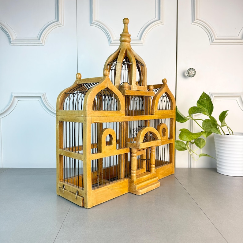 May include: A wooden birdcage with a domed top and intricate details. The cage is painted in a light brown color and has a wire mesh interior. The cage is sitting on a gray surface.