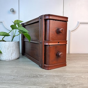 May include: A small, two-drawer wooden chest with a dark reddish-brown finish. The drawers have round wooden knobs. The chest is framed with darker wood and metal rivets. A potted plant with green leaves sits to the left.