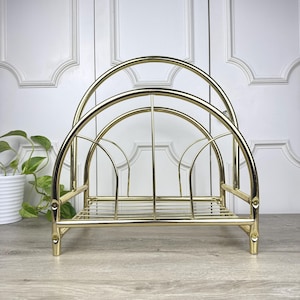 May include: A gold-colored metal magazine rack with a curved, arched design. The rack has a rectangular base and vertical bars for holding magazines or papers. The rack is made of thin metal rods and has a shiny finish.
