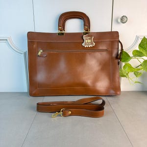 May include: A brown leather briefcase with a gold handle and a detachable shoulder strap. The briefcase has a zippered pocket on the front and a label that says "Italy Leather".