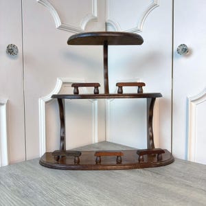 May include: A dark brown, three-tiered corner shelf with a semi-circular base and a rounded top shelf. The shelf has a central support and several small, rectangular pegs for holding items. The wood has a rich, warm tone.
