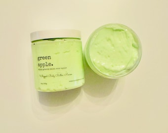 May include: Two containers of green apple whipped body butter mousse. One jar is closed with a white lid, and the other is open, revealing the fluffy, light green product. The label reads "green apple" and "crisp granny smith sour apple."