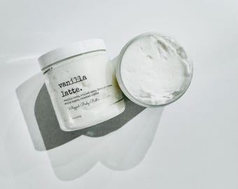 May include: Two white jars of whipped body butter, one with the lid on and the other open, revealing the creamy contents. The jar on the left has the text "vanilla latte." printed on it. The ingredients include vanilla beans, rolled oats, and roasted coffee.