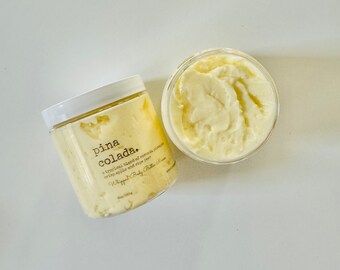 May include: A white jar with a yellow lid filled with a whipped body butter. The jar has a black label with the text "pina colada."  A tropical blend of coconut, crisp apple and ripe pear. Whipped Body Butter. 8oz/226g.