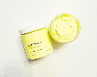 May include: A yellow foaming sugar scrub in a clear jar with a white lid. The jar is labeled "gardenia.  orchid + rose + orange  foaming sugar scrub".