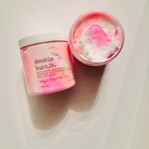 May include: Two containers of whipped body butter. One is open, revealing pink and white swirls. The other is closed, with the text "double bubble" and product details. The product is described as having a strawberry, sunburst orange, and mint scent.