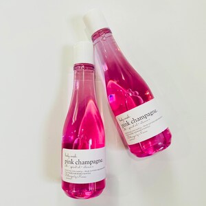 May include: Two pink glass bottles of body wash with white caps. The bottles are labeled "pink champagne" and feature a pink and white design. The bottles are on a white background.