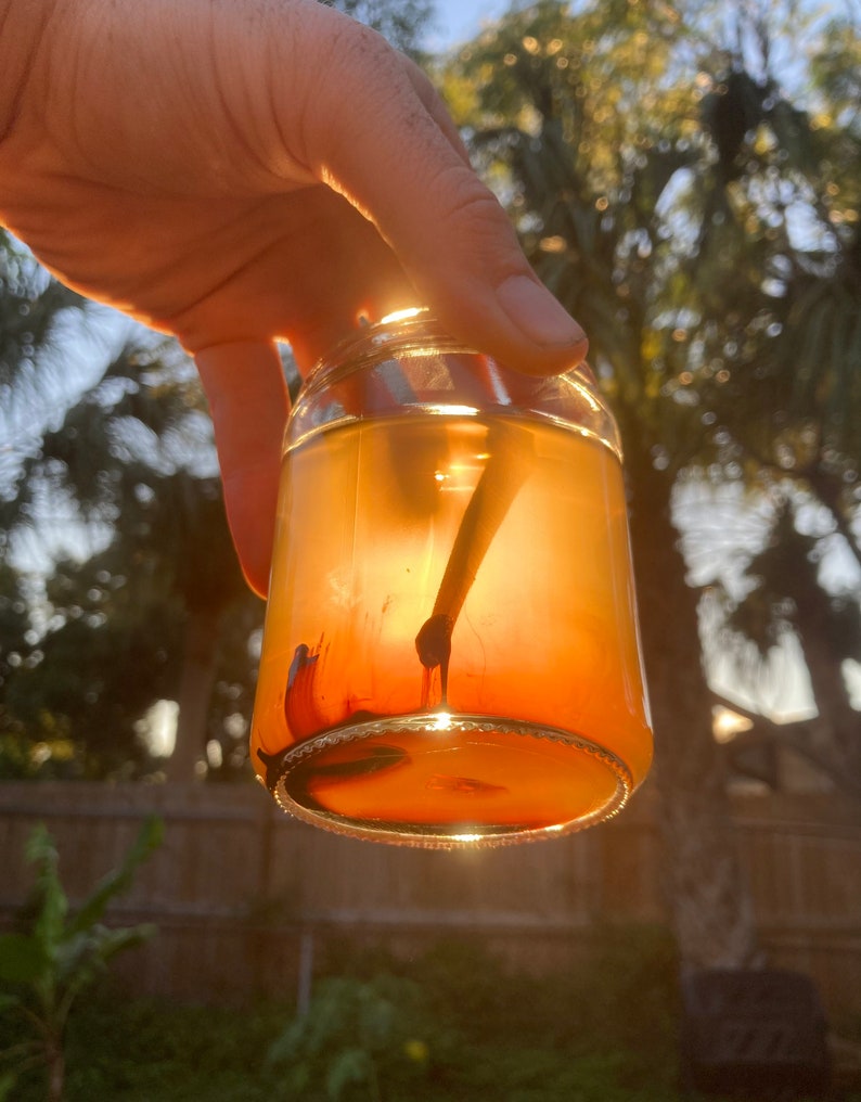 May include: A clear glass jar with a red liquid inside. The jar is being held by a hand and the sun is shining through the liquid, creating a bright orange glow.