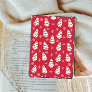 May include: A red tablet case featuring a repeating pattern of white Christmas tree cookies and snowflakes. The cookies have white icing and are decorated with small, dark sprinkles. The case is held by a person wearing a cream-colored sweater.