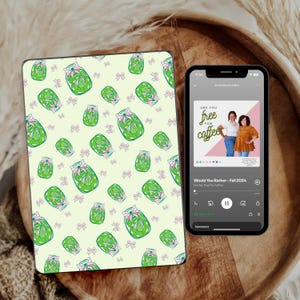 May include: A digital tablet with a green and pink pattern of jars with green pickles and pink bows. The tablet is next to a smartphone with a podcast app open. The podcast is called "Would You Rather - Fall 2024" and the episode is called "Are You Free For Coffee?"