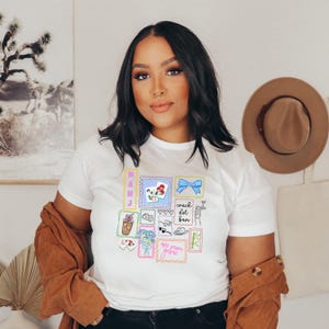 May include: White t-shirt with a colorful, illustrated design of postage stamps. The stamps feature various images, including flowers, a bow, and text. The shirt has short sleeves and a crew neck.