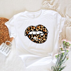 Puede incluir: Camiseta blanca con un gráfico de una boca de estampado de leopardo mordiendo un labio. El gráfico es en marrón, negro y blanco.