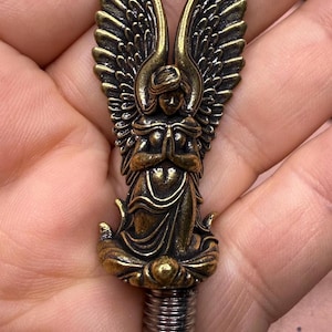 May include: A bronze-colored angel figurine with outstretched wings and clasped hands. The angel is kneeling on a decorative base, and the bottom has a threaded metal screw. The wings have detailed feathering.